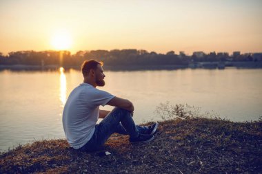 Young handsome bearded caucasian blond man sitting on cliff and looking at sunset. In background is river. clipart