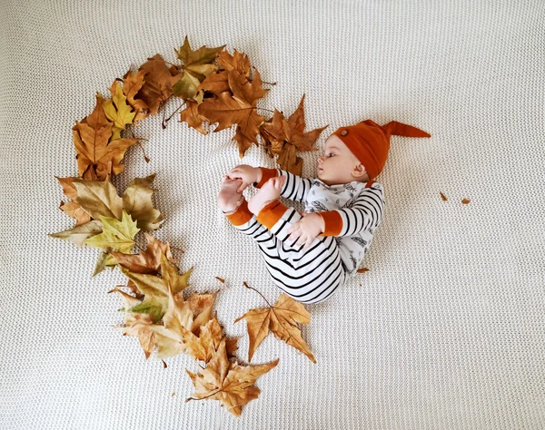 可愛い赤ちゃん服を着た可愛い赤ちゃんのトップビューがベッドの上に横たわり 秋の葉でハートの形を作っています — ストック写真