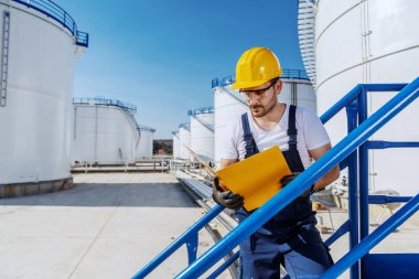Hardworking handsome caucasian worker in overall and with helmet on head standing on the stairs and looking at folder with documents. Oil production. clipart