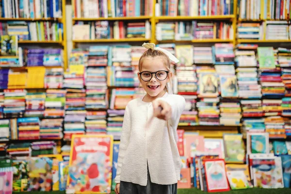 Adorable smiling little caucasian girl with eyeglasses and ponytail pointing with sword toy and looking at camera while standing in bookstore.