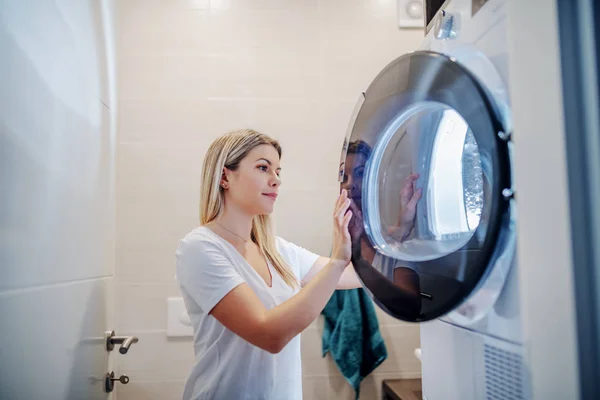 Worthy caucasian young blond housewife closing drying machine while standing in bathroom in late hours.