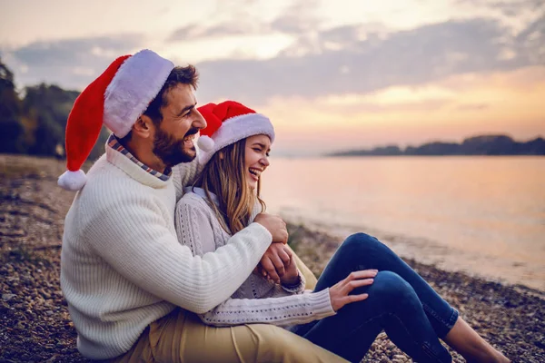 美しい白人のカップルの愛の海岸の川と抱擁の横に座っている どちらも白いセーターに身を包み サンタの帽子を頭にかぶっています クリスマス タイム — ストック写真
