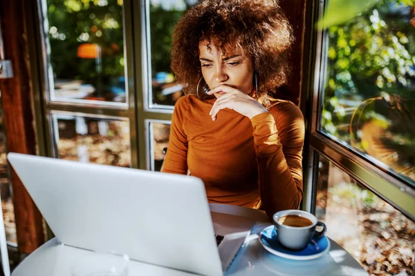 巻き毛の可愛い若い混合人種の女性と カフェに座ってノートパソコンを見ているタートルネック テーブルの上には新鮮な朝のコーヒーのカップ — ストック写真