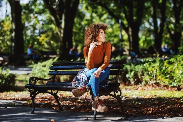 年轻迷人的 面带微笑的混血女子 穿着高领毛衣 卷曲的头发 坐在公园的长椅上 用智能手机交谈 — 图库照片