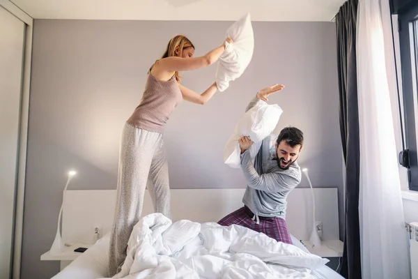 若い魅力的な白人のカップルは 午前中に寝室で枕の戦いをしています 二人ともパジャマ姿 — ストック写真
