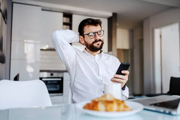若い白人の重大なビジネスマンで眼鏡をかけてダイニングルームのテーブルに座り スマートフォンでニュースを読んでいます テーブルには朝食とラップトップがあります — ストック写真