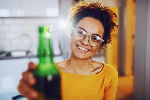 Cheerful smiling cute tanned caucasian brunette with curly hair sitting at party and toasting with beer.