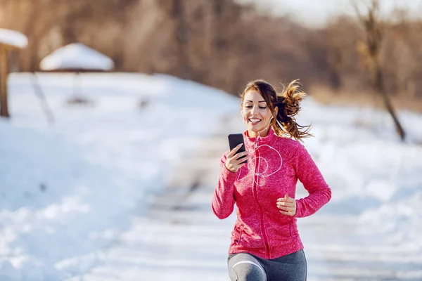 Smiling caucasian brunette in sportswear and with earphones in ears looking at smart phone while running in nature. Wintertime. Outdoor fitness concept.