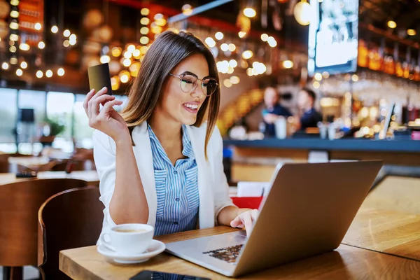 迷人的 时髦的 面带微笑的高加索黑发女人坐在咖啡店里 拿着信用卡 用笔记本电脑上网搜索 在商店购物后 她决定在网上购买更多的商品 — 图库照片