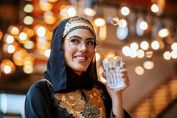穿着传统服装 面带笑容的迷人的积极的穆斯林妇女独自坐在咖啡店里 举着一杯水 — 图库照片