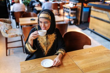Gorgeous smiling positive arab woman in traditional wear sitting in coffee shop alone and enjoying her fresh morning coffee. clipart