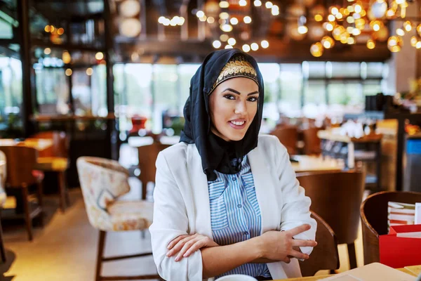 魅力的な現代的な笑顔イスラム教徒の女性とスカーフでカフェテリアに座って手を折り畳んでカメラを見て — ストック写真
