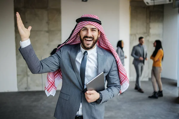 Overjoyed arab rich investor in suit standing in building in construction process, holding tablet under armpit and feeling happy to invest in building on such a good location.