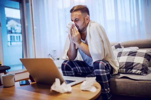 穿着睡衣 头戴毛毯的英俊 不修边幅的白人男子坐在客厅的沙发上 吹着鼻涕 桌上有笔记本电脑 茶杯和用过的手帕 — 图库照片