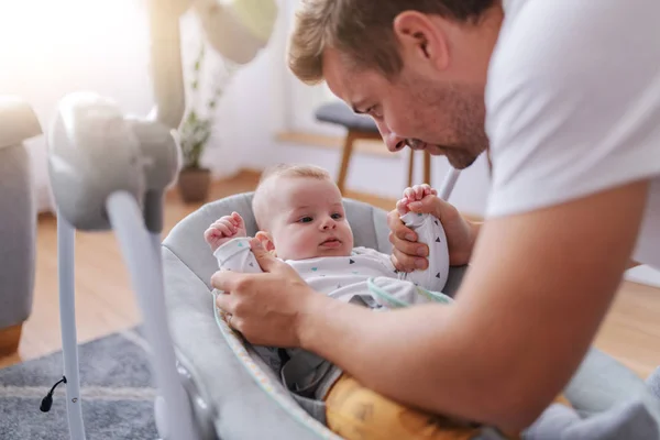 愛らしい6ヶ月の息子と遊ぶハンサムな思いやりのある白人の若い父親 赤ちゃんはベビーロッカーチェアに横になり 満足感 — ストック写真