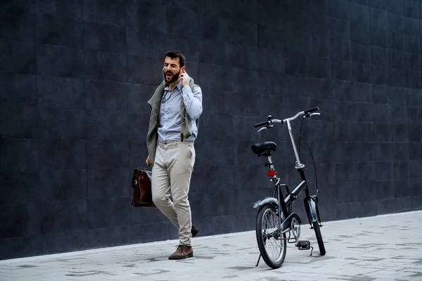 轻松愉快的英俊时尚的高加索商人一边工作一边听音乐 唱歌和提着公文包 他旁边是自行车 背景是灰墙 — 图库照片