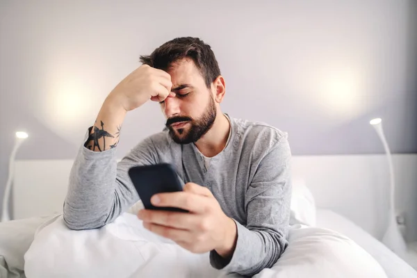 Depressed lonely caucasian bearded man sitting on bed in bedroom at night, holding his head and using smart phone for online chatting. Problems in relationship.