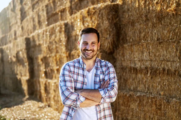 Handsome caucasian smiling farmer standing outdoors with arms crossed and looking at camera. In background are of hay.