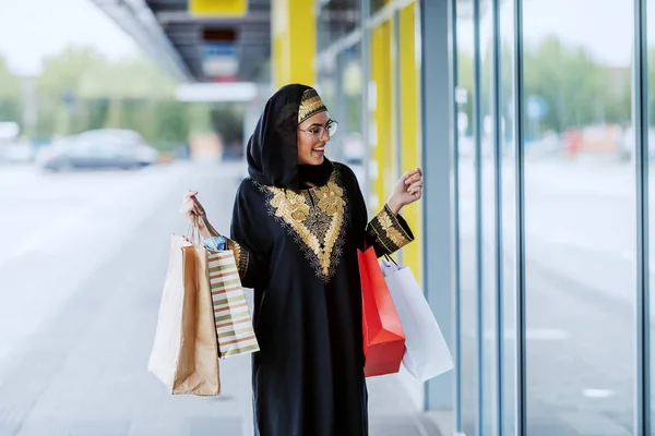穿着传统服装的美丽的穆斯林妇女惊奇地站在户外 手里拿着购物袋 望着商店橱窗 她想在特殊的场合买件漂亮的衣服 — 图库照片
