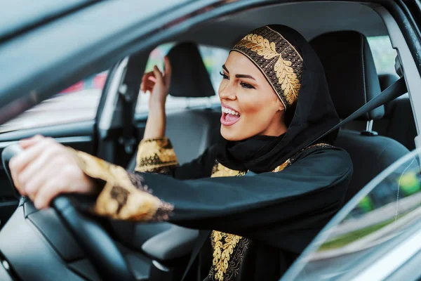 穿着传统服装 开新车 听音乐 唱歌的快乐而华丽的积极的穆斯林女性 — 图库照片