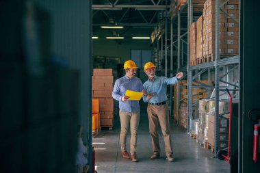 CEO going around warehouse with supervisor and talking analyzing sale statistics. Younger man holding folder with data while older one holding tablet and pointing at boxes. Both having yellow helmets. clipart