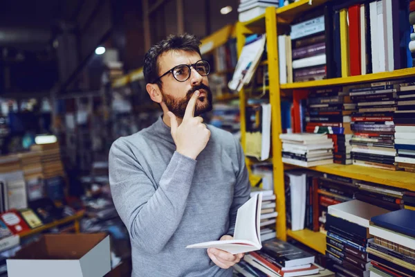魅力的なCaucasian深刻なひげを生やした男で眼鏡を立って書店で手に本と思考 — ストック写真
