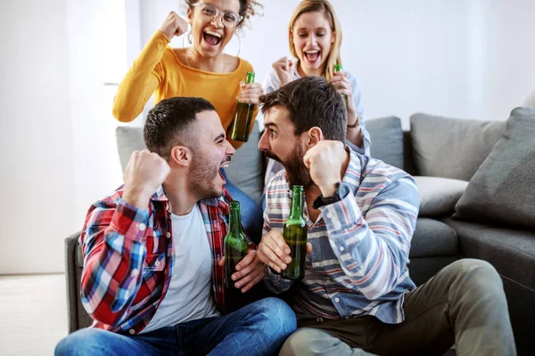 Group of friend sitting in living room and cheering for their favorite football team. They all holding beer bottles.