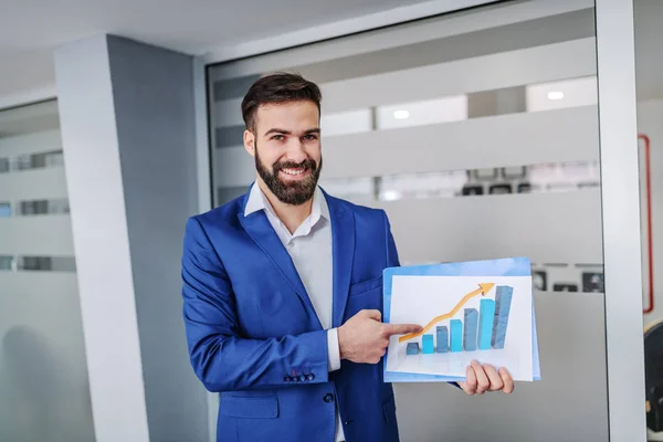 Young successful smiling broker standing in hall of his firm, holding chart and pointing at it. Stocks on market going up.