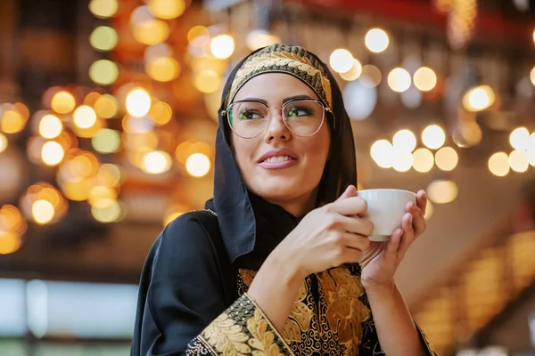 穿着传统服装的迷人而积极的穆斯林妇女独自坐在咖啡店里 举着一杯新鲜咖啡 望着远方 — 图库照片