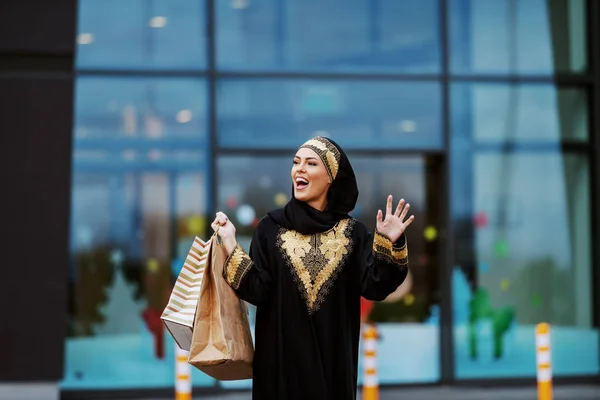 手にショッピングバッグを持つショッピングモールの前に立つ伝統的な服を着た豪華な肯定的な笑顔のイスラム教徒の女性は友人に手を振って — ストック写真
