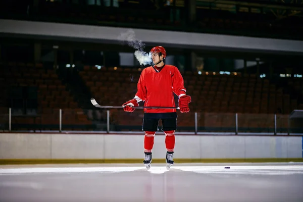 Tired hockey player standing on ice with stick in hands in hall.