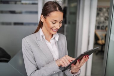 Portrait of hardworking positive Caucasian young businesswoman standing inside corporate firm and using tablet for reading an e-mail. clipart