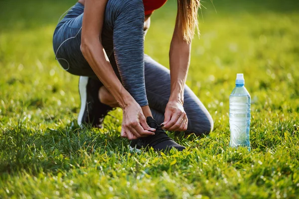 穿着运动服的高加索女人跪在草地上 鞋带绑在运动鞋上的剪影 她旁边是一瓶水 大自然阳光灿烂的早晨 — 图库照片