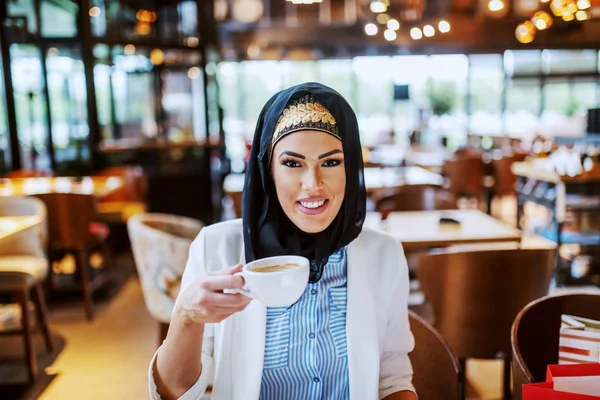 迷人的现代微笑的穆斯林女人坐在咖啡馆里 举着一杯咖啡 坐在她旁边的是购物袋 她整天都在买东西 现在休息一下 — 图库照片