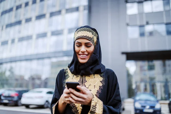 Piękna Uśmiechnięta Muzułmanka Stojąca Przed Budynkiem Korporacji Używająca Smartfona Wysyłania — Zdjęcie stockowe