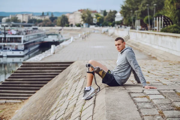 Side view of caucasian handicapped sportsman in sportswear and with artificial leg sitting on quay and looking away.