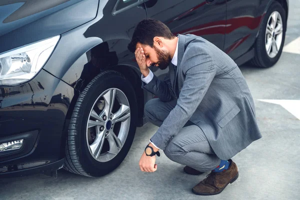Nervous bearded businessman crouching next to his car and holding head. Tire is flat. How to get on time to work now?