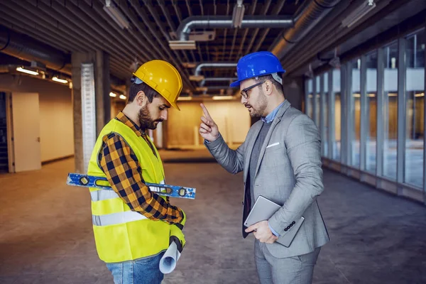 Angry caucasian businessman in suit and helmet on head arguing with irresponsible construction worker. Building in construction process interior.