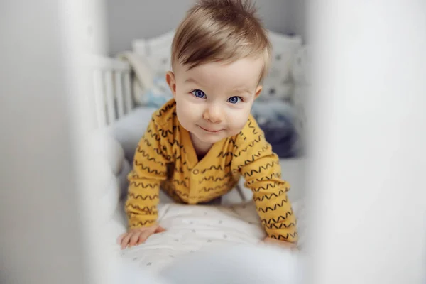 美しい青い目をした愛らしい小さな金髪の赤ん坊の男の子は 笑みを浮かべて 耳のカメラを見て ベビーベッドでクロールします 朝の時間 — ストック写真