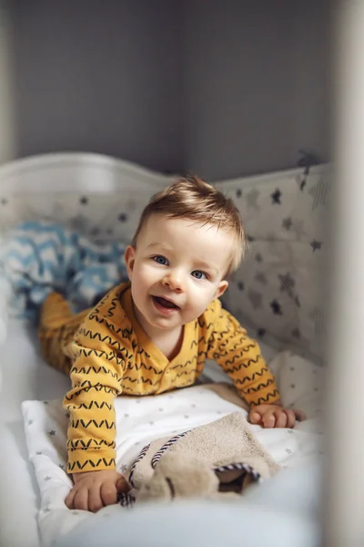 美しい青い目をした魅力的な笑顔のブロンドの赤ん坊の少年は 彼のカニの胃の上に横たわって カメラを見ています 朝の時間 — ストック写真