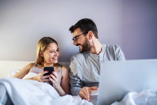 Jovem Casal Feliz Sentado Cama Manhã Usando Laptop Telefone Inteligente — Fotografia de Stock