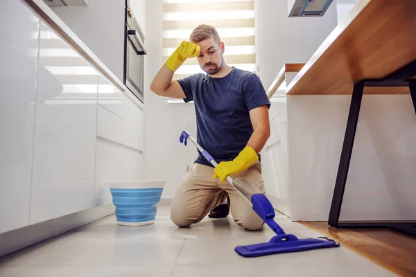 Young tidy worthy tired bearded man kneeling, wiping sweat and trying to clean dirty floor in kitchen.