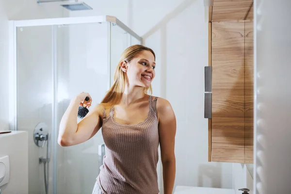 鏡の前のバスルームに立つ若い笑顔のブロンドと彼女の髪を持っています 朝のルーチン — ストック写真