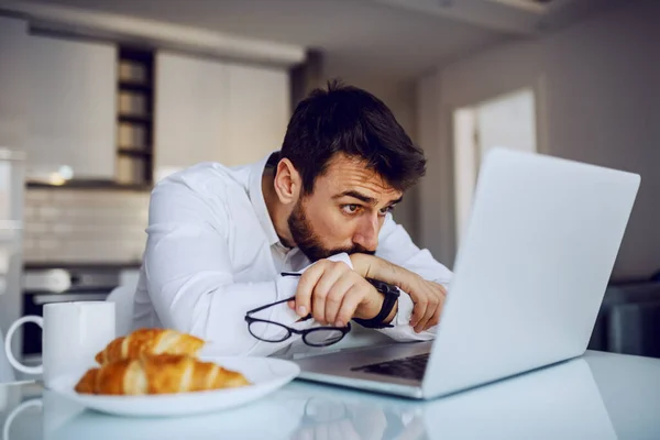 年轻英俊的大胡子男子坐在餐桌前 靠在桌子上 看老板用笔记本电脑发来的电子邮件 脸上露出严肃的表情 — 图库照片