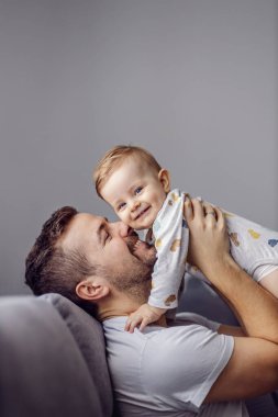 Young good-looking father holding his beloved little son and playing with him while sitting in living room. Toddler is looking at camera and smiling. clipart