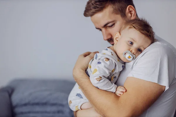 Sleepy adorable little boy with beautiful blue eyes having pacifier in his mouth and leaning on father\'s shoulder. Father is holding his beloved son and putting him to sleep. Nap time.