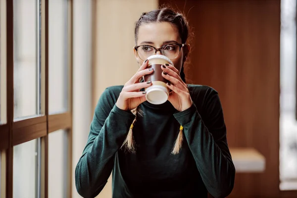 窓の近くの喫茶店に座っている若い女性と使い捨てのカップから新鮮なコーヒーを飲む — ストック写真