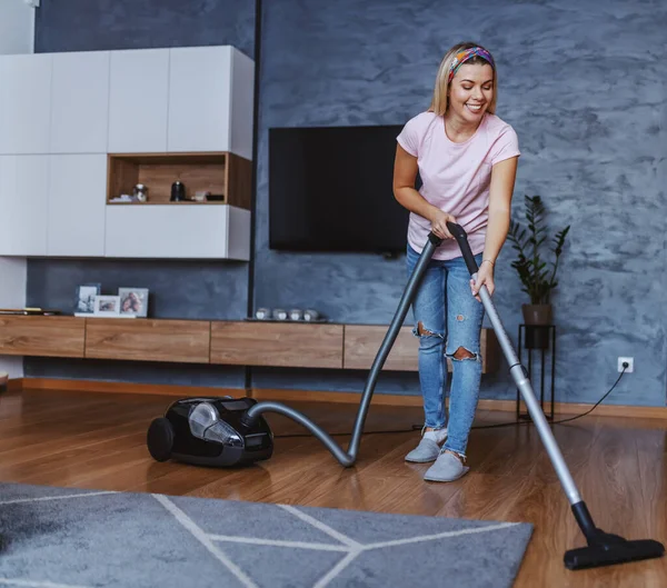 Beautiful tidy caucasian blond young woman using vacuum cleaner to clean living room floor.