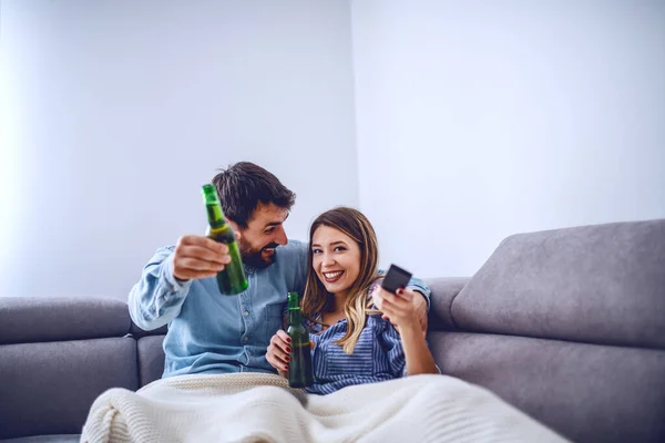 リビングルームにある毛布で覆われた魅力的な白人のカップルは テレビを見てビールを飲んでいます リモコンとチャンネルを変える女 — ストック写真