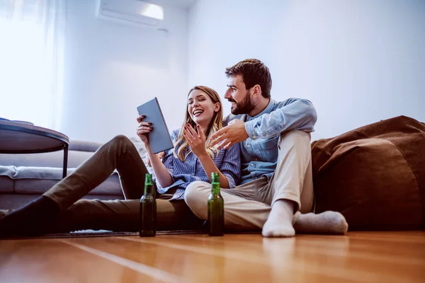 リビングルームで床に座ってタブレットを見て愛の若い陽気な白人のカップル 床にはビールボトルが置かれている 日曜日の午後の無料時間 — ストック写真